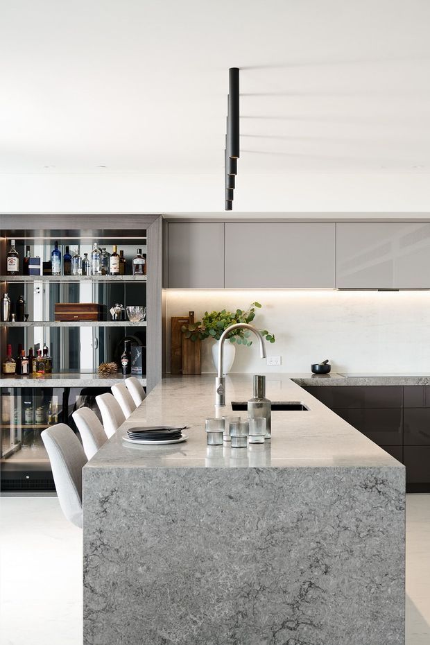A bright contemporary kitchen centered on a large grey stone island with an integrated sink, sculptural tap, seating for four, and pendant lighting. Flat-panel grey cabinetry, a pale illuminated splashback, dark lower cabinets, and a glazed built-in drinks display with bottles and glassware complete the refined minimalist fit-out.
