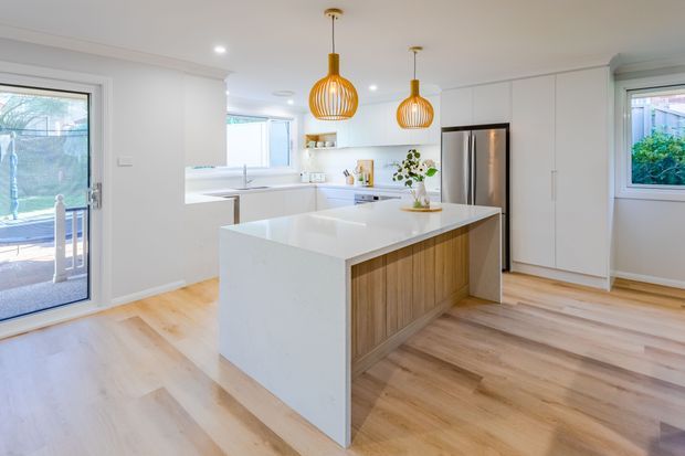 Bright contemporary kitchen featuring a large white waterfall island with a timber-fronted base, white cabinetry, stainless-steel refrigerator, light timber flooring, expansive windows and two woven pendant lights. The space is open, clean and softly warm in tone.