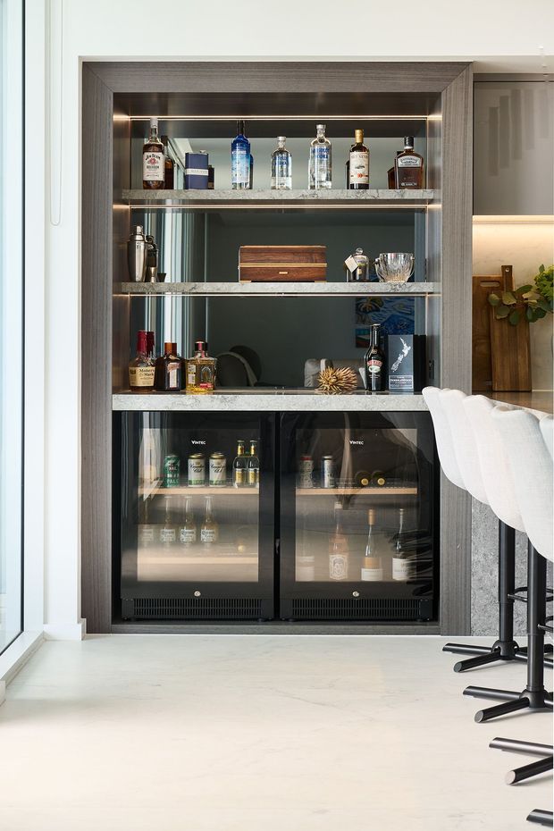 A built-in home bar integrated into a modern kitchen or entertaining area. The dark timber-framed niche has illuminated glass shelves displaying bottles and barware, with two under-counter glass-door beverage refrigerators below. Pale stone flooring, a light stone counter edge, black metal bar stools, and adjacent cabinetry create a refined contemporary setting.