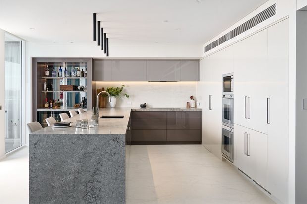 Contemporary open-plan kitchen with a large grey stone island, integrated dark cabinetry, white floor-to-ceiling cabinetry, built-in appliances, pale stone splashback, and a slim linear pendant light. A dining area and open shelving with bottles are visible to the left.