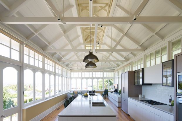 A bright, elongated kitchen and dining space within a glazed, pavilion-like room. White-painted exposed roof trusses and beams create a high pitched ceiling, while extensive arched and rectangular windows line both sides. The kitchen features a long white island with integrated sink, pale cabinetry, a dark stone-look benchtop, and suspended industrial-style pendant lights. Timber flooring and abundant daylight give the space an airy, refined character.