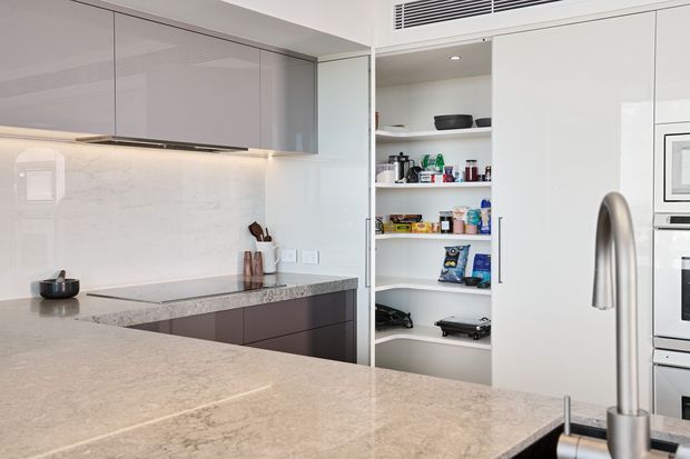 Contemporary minimalist kitchen featuring pale stone-look benchtops and splashback, glossy light-grey overhead cabinetry, dark lower cabinetry, integrated appliances, a concealed walk-in pantry with open shelving, and a sculptural stainless-steel tap at the island sink.