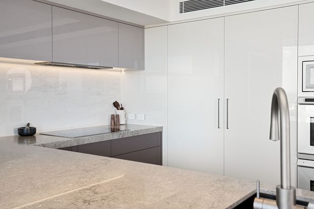 A sleek contemporary kitchen with a large pale stone island and integrated sink, handleless grey and white cabinetry, a pale marble-look splashback, built-in stainless-steel appliances, and a curved brushed-metal tap. The space is clean-lined and softly lit with a neutral palette.