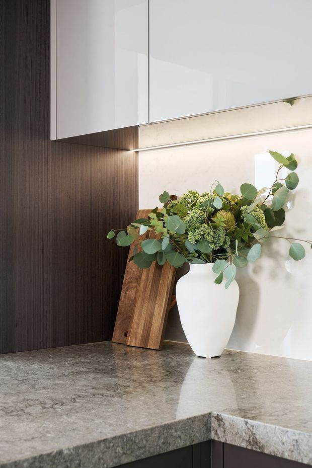 Close-up view of a refined kitchen corner featuring dark vertically grained cabinetry, glossy white overhead cabinets, a pale stone or marble splashback, and a substantial grey veined stone benchtop. Warm integrated under-cabinet lighting highlights a white sculptural vase with leafy greenery and a timber chopping board leaning against the wall.