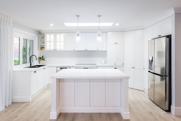 Bright, spacious white kitchen with a large paneled island, shaker-style cabinetry, stainless-steel appliances, pale timber-look flooring, pendant lights, and abundant natural light from windows and a skylight.