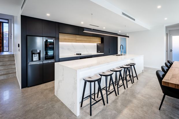 A spacious contemporary kitchen with a long white stone island and seating for six, matte black full-height cabinetry, integrated stainless-steel appliances, a marble-look splashback, warm timber open shelving, and concrete-look flooring. A dining area is partially visible at right, with stairs and a hallway beyond.