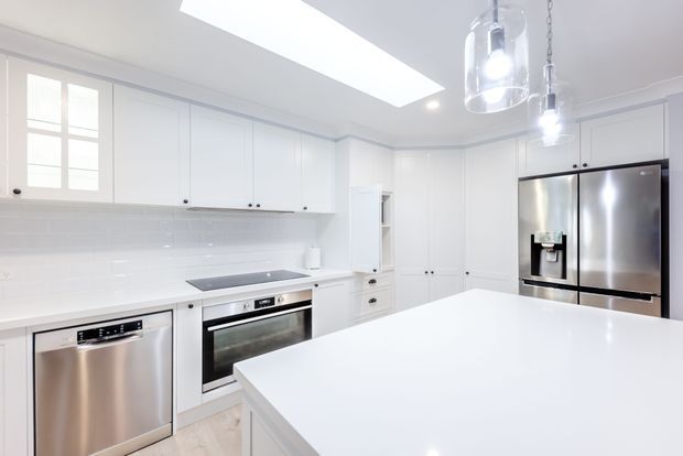 Bright contemporary white kitchen with extensive floor-to-ceiling cabinetry, a large white island, integrated stainless-steel oven and dishwasher, and a stainless-steel French-door refrigerator. White subway-tile splashback, pale flooring, pendant lights, and a large skylight create a clean, airy interior.