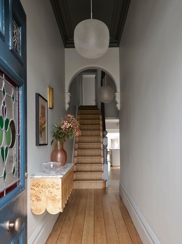 A narrow, refined residential entry hall viewed from the front door, featuring a timber staircase with woven runner, arched opening, ornate ceiling mouldings painted dark charcoal, stained-glass entry door, marble-topped timber console, decorative wall lighting, pendant globe lights, and warm timber flooring. The hall leads toward a brighter living area beyond.