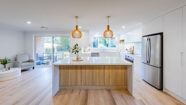 Bright open-plan kitchen with a large central island featuring a light timber-panelled base and pale stone benchtop. White cabinetry lines the rear wall, with a stainless-steel French-door refrigerator, integrated appliances, and a window above the sink. Two warm pendant lights hang over the island, while light timber flooring continues into the adjacent living area with sliding glass doors and an armchair.
