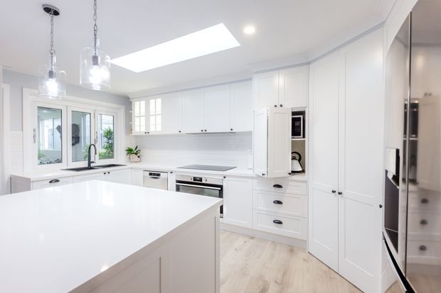 Bright, spacious white kitchen with extensive shaker-style cabinetry, a large white island, integrated appliances, a skylight, pendant lights, and pale timber-look flooring. The design is clean and refined with abundant natural light.