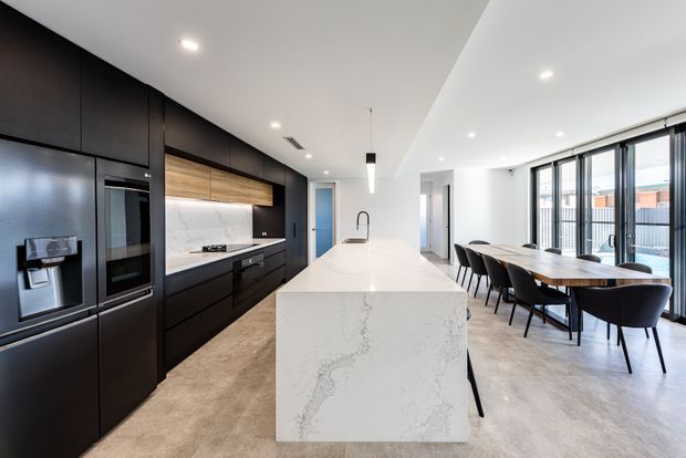 Contemporary open-plan kitchen and dining area with a large white stone island, matte black full-height cabinetry, integrated appliances, timber accent joinery, and a long dining table beside floor-to-ceiling glazed doors. Recessed lighting and a clean, linear layout create a bright, refined interior.