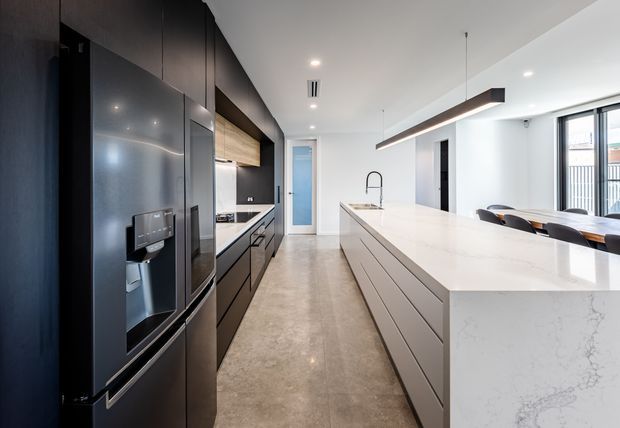 Contemporary galley-style kitchen with a large white stone island and integrated sink, dark full-height cabinetry, a stainless-steel refrigerator, warm timber cabinetry accents, polished concrete-look flooring, and linear pendant lighting. A dining area and bright glazing are visible beyond the island.