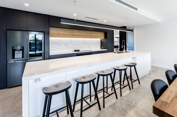 Contemporary open-plan kitchen with a large white island and five black timber bar stools, dark floor-to-ceiling cabinetry, integrated appliances, a pale marble-look splashback, warm timber overhead cabinetry, and a timber dining table at right. The space has a clean, refined palette with linear pendant lighting and recessed ceiling lights.