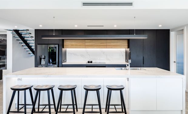 Contemporary open-plan kitchen dominated by a large white island with five black stools. The kitchen features matte black full-height cabinetry, warm timber overhead cabinetry, a white marble-look splashback, integrated appliances, and a long linear pendant light. A floating open-riser stairway is visible at the left, with bright white walls and ceiling throughout.