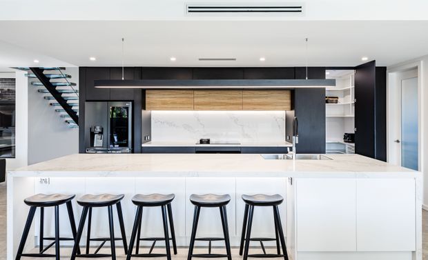 Bright contemporary kitchen dominated by a large white island with five black bar stools. The kitchen features matte dark cabinetry, warm timber overhead cupboards, a white marble-look splashback, integrated appliances, open shelving, and a minimalist linear pendant light. A sculptural open-riser stair is visible at left, with a hallway or doorway at right.