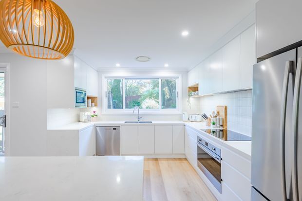 Bright contemporary white kitchen with an L-shaped layout and large central island. The room features white flat-panel cabinetry, pale timber flooring, a white benchtop, tiled splashback, stainless-steel appliances, a large window overlooking greenery, and a prominent woven timber pendant light.