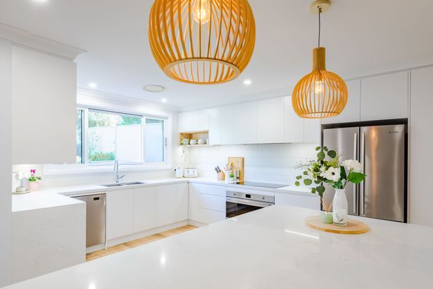 Bright, spacious white kitchen with an L-shaped run of cabinetry, a large central island, stainless-steel appliances, white tiled splashback, and pale timber flooring. Two oversized woven pendant lights provide warm natural texture above the island, while a flower arrangement adds a small decorative accent.