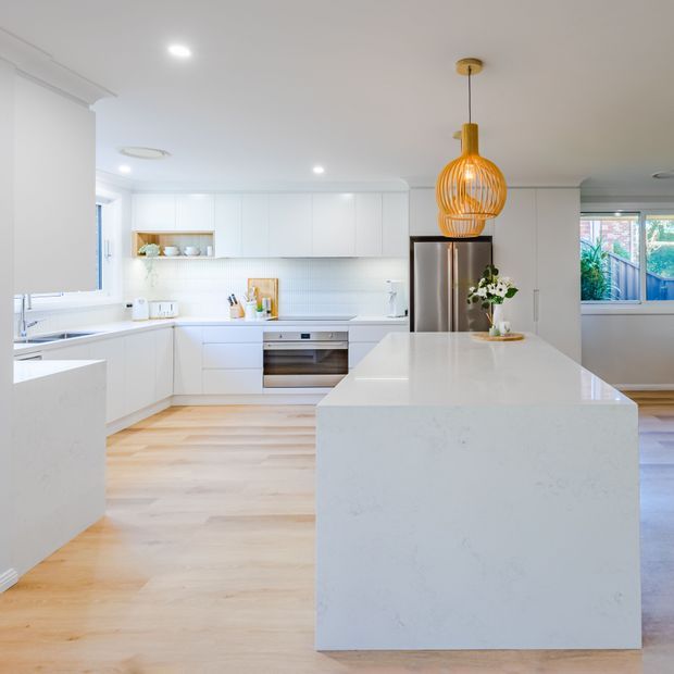 Bright contemporary white kitchen with extensive built-in cabinetry, a large white stone island, integrated stainless-steel appliances, pale timber flooring, and a sculptural warm-toned pendant light. The open-plan space is naturally lit through windows and has a clean, spacious feel.