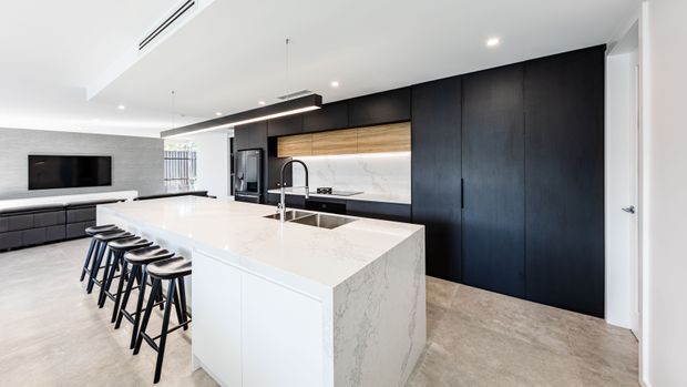 Contemporary open-plan kitchen with a large white stone island and integrated sink, seating for several people, dark full-height cabinetry, a timber feature cabinet section, and a light marble-look splashback. The kitchen opens to a living area with wall-mounted television and built-in cabinetry. Recessed lighting and linear pendant fixtures create a clean, refined interior.