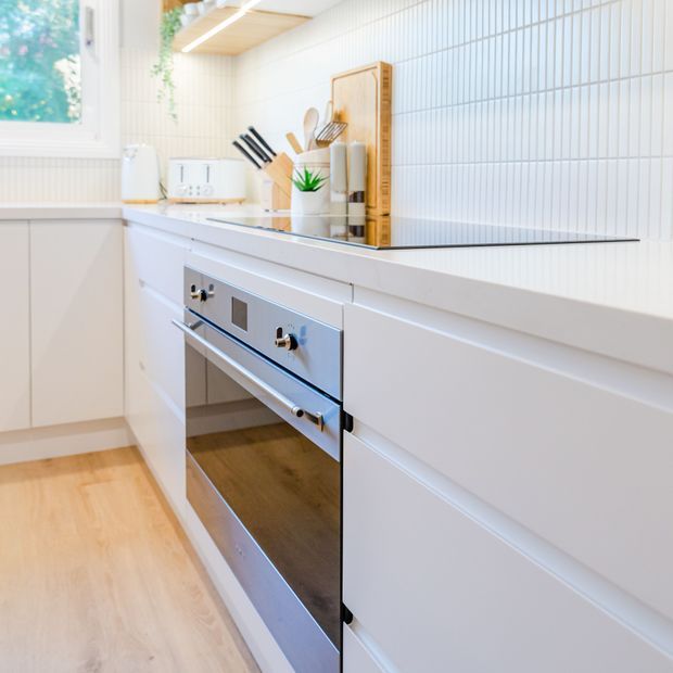 Bright, compact contemporary kitchen focused on white built-in cabinetry, a stainless-steel oven, pale timber flooring, and a white vertically ribbed tiled splashback. The benchtop holds a cutting board, knife block, utensils, and small appliances, while daylight enters through the window.