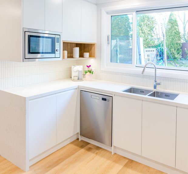 Bright, compact contemporary kitchen with white flat-panel cabinetry, integrated microwave, stainless-steel dishwasher and double-bowl sink beneath a large window. The kitchen features a white tiled splashback, pale engineered-looking benchtops, light timber flooring, and a small open timber display niche. Natural light and views to greenery make the space feel clean and airy.