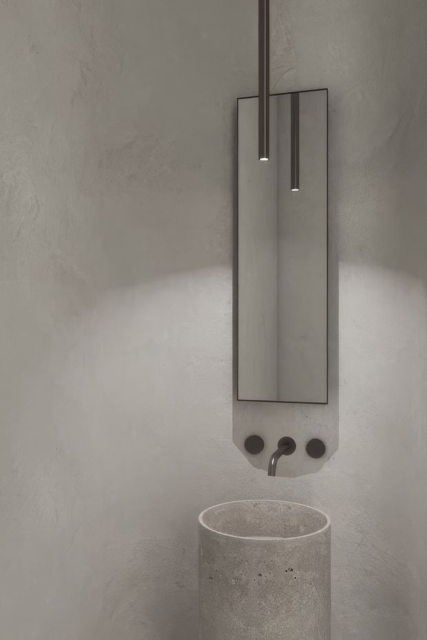 A minimalist bathroom vanity vignette with a cylindrical concrete basin, wall-mounted dark tapware, a tall rectangular mirror, and two slim pendant lights. The walls have a textured grey plaster or concrete-like finish, creating a monochromatic, sculptural interior.