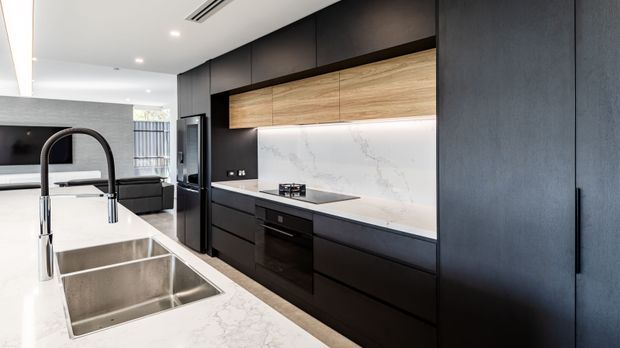 Contemporary open-plan kitchen with a large white stone island, double stainless-steel sink, matte black cabinetry, warm timber overhead cabinetry, and a white veined stone splashback and benchtop. The kitchen opens toward a living area in the background.