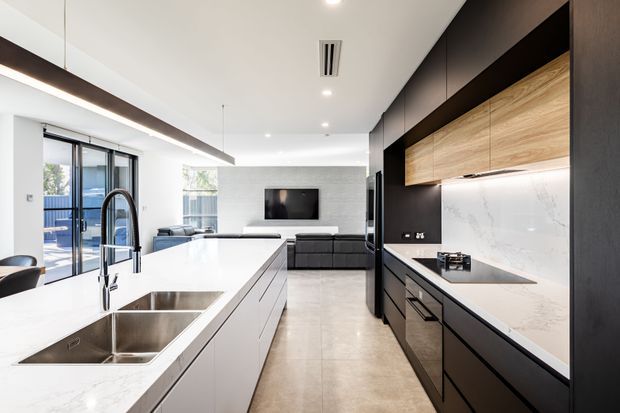 A bright contemporary open-plan kitchen with a large white stone island, double stainless-steel sink and curved black tap. Matte black floor-to-ceiling cabinetry lines the right wall, paired with warm timber overhead cupboards and a pale stone splashback and benchtop. The kitchen opens toward a living area with a dark sofa and wall-mounted television, with generous glazing and recessed lighting creating an airy, refined interior.