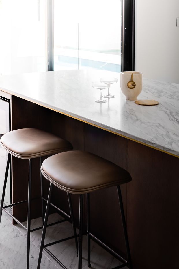 Close-up of a refined kitchen island or breakfast bar with a white veined stone benchtop, dark timber cabinetry, and a slim brass-gold edge detail. Two upholstered brown bar stools sit beneath the overhang, with floor-to-ceiling glazing behind and minimalist decorative objects on the counter.