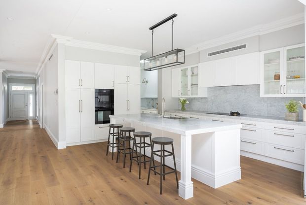 Bright, spacious white kitchen with a large central island and seating for five, extensive white cabinetry, integrated appliances, glass-front upper cabinets, and a grey stone backsplash and benchtop. The room has warm timber flooring, classic wall mouldings, a linear black-and-glass pendant light, and opens to a hallway.