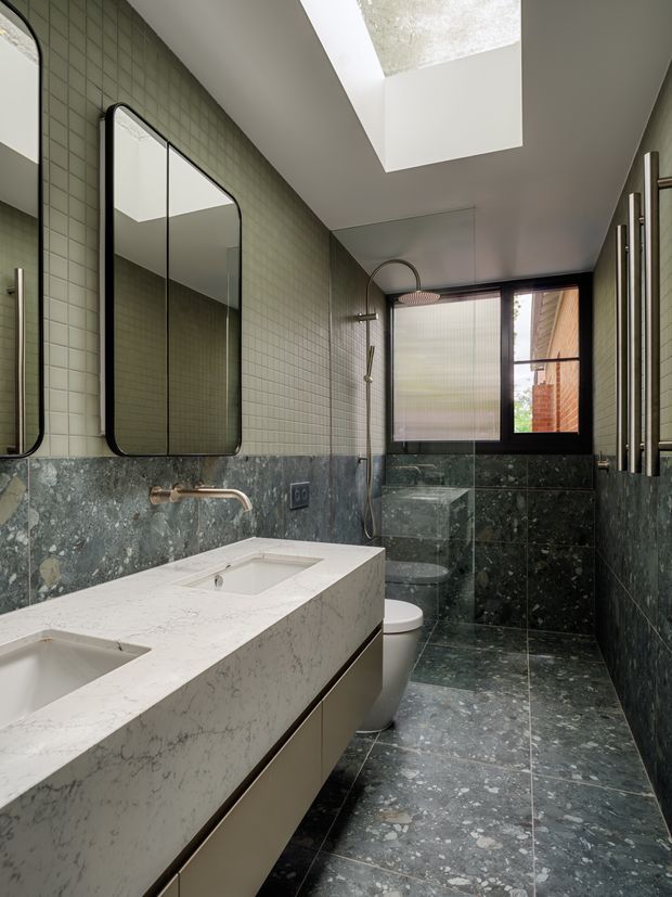 A contemporary bathroom with a double vanity, pale stone benchtop, dark green stone wall and floor tiles, small sage-green wall tiles, rounded black-framed mirrors, brass tapware, a wall-mounted shower, toilet, and a large skylight. The space has a refined, moody palette with natural stone textures and strong vertical proportions.