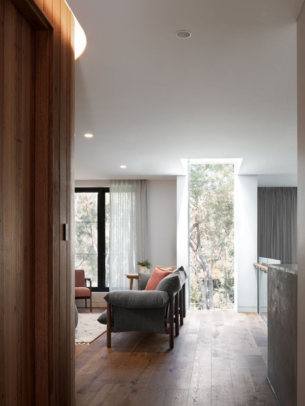 A bright, contemporary open-plan living area viewed from a timber-lined entry or hallway. The room features wide timber flooring, a grey upholstered lounge chair and coral accent cushion, sheer curtains, black-framed glazing, and a dramatic tall vertical window bringing in views of trees. A compact kitchen with dark grey stone-look cabinetry or benchtop is visible on the right. The space has a clean white ceiling with recessed lighting and warm natural materials.