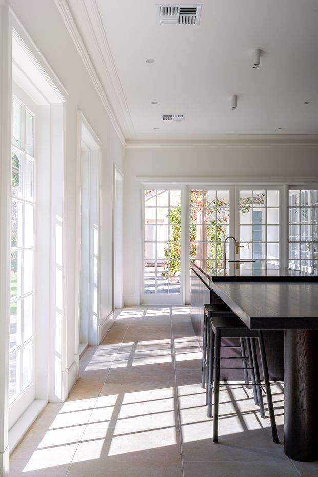 A bright, refined kitchen with extensive white-framed glazed doors and windows, classical wall and ceiling mouldings, pale large-format tiled flooring, and a large dark island with integrated seating. Strong sunlight creates geometric shadows across the floor, while a leafy courtyard or garden is visible beyond the glazing.
