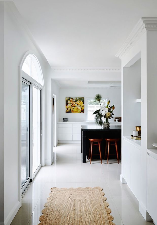 Bright, predominantly white open-plan kitchen interior with tall arched glazing and expansive glass doors on the left, glossy light tiled flooring, decorative ceiling cornices, and white built-in cabinetry. A contrasting black kitchen island with timber bar stools forms the focal point, complemented by a floral arrangement and colourful wall artwork. The space has a refined, airy feel with classic architectural detailing and contemporary joinery.