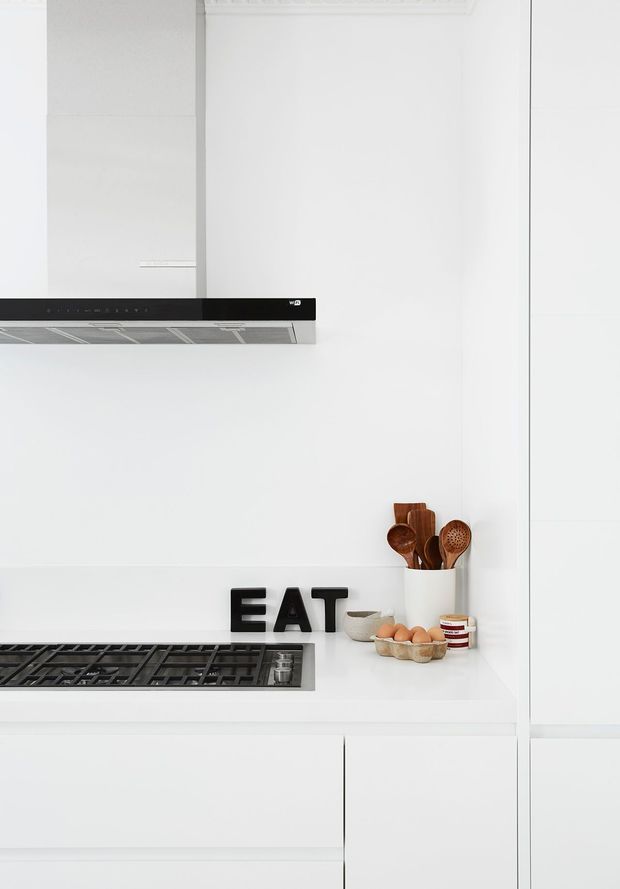 A bright, minimalist white kitchen with seamless white cabinetry and benchtops, an integrated black gas cooktop and matching rangehood, and a white splashback. Wooden cooking utensils in a white container, eggs, bowls, and small pantry jars add warm accents. Decorative black 'EAT' lettering sits on the benchtop.