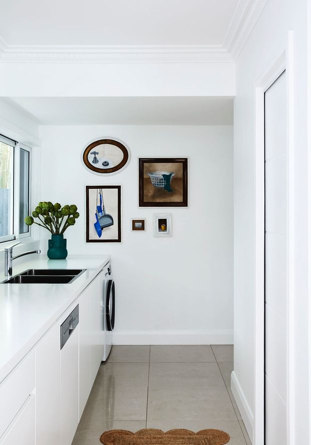 Bright, compact white utility room or laundry with a long white benchtop, integrated stainless-steel sink and tap, white cabinetry, a front-loading washing machine, tiled floor, and a window above the work area. Decorative framed artworks and a vase add character, while ornate ceiling mouldings and crisp white trim give the room a traditional refined feel.