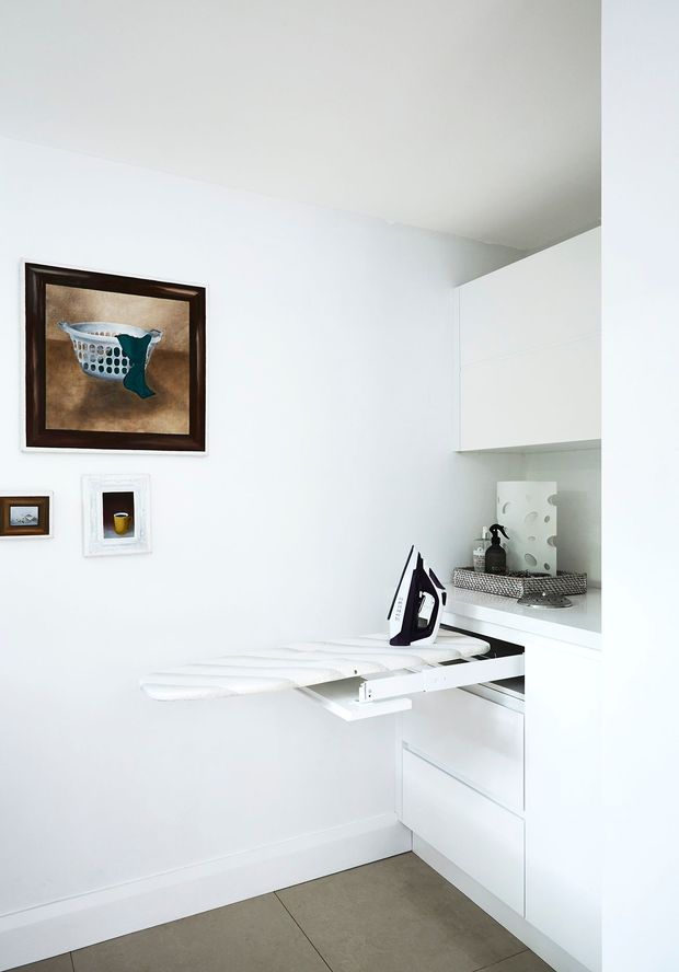 Compact, bright white laundry room with built-in cabinetry, an integrated pull-out ironing board, iron, open shelving, and a tiled floor. Framed artwork adds a small decorative touch to the otherwise minimalist utility space.