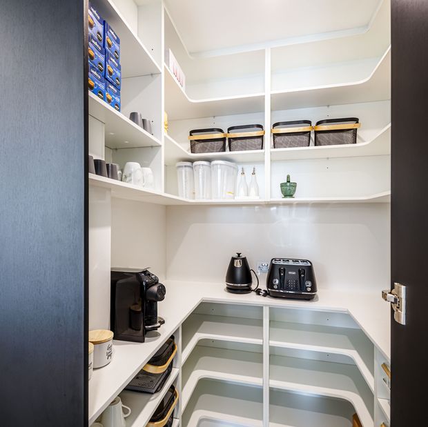 A compact, highly organised walk-in pantry with white built-in open shelving, curved shelf edges, integrated corner storage, a light benchtop, and a glossy pale splashback. Shelves hold baskets, cups, containers, a coffee machine, kettle, toaster, and other small appliances. The space is bright and clean, viewed through an open dark-framed door.