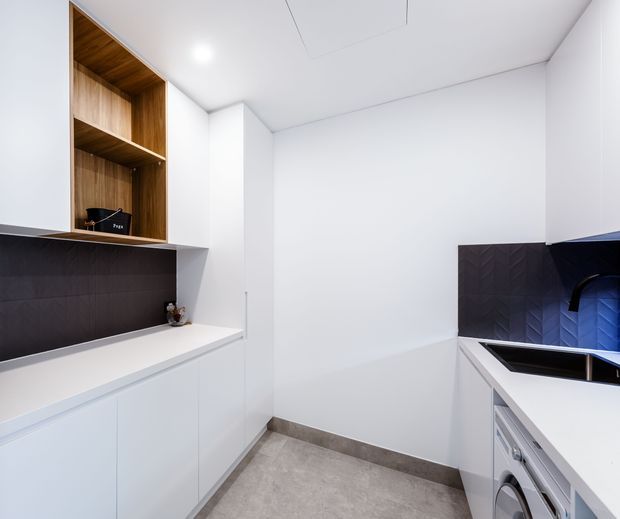 Bright, compact contemporary laundry with white built-in cabinetry and benchtops, dark textured splashback panels, an open timber-lined overhead niche, integrated washing machine, and a deep sink. The space has a clean, minimalist fit-out with grey tiled flooring and recessed ceiling lighting.