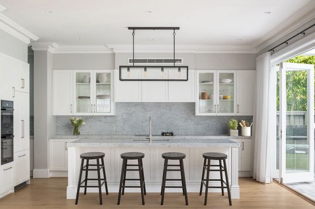 Bright, spacious contemporary kitchen with white shaker-style cabinetry, glass-front display cupboards, a full-height grey stone backsplash, and a matching island with seating for four. Dark timber bar stools and black linear pendant lighting provide contrast, while bi-fold or hinged glazing opens to an outdoor area. The room has light timber flooring, white walls and ceiling detailing, and abundant natural light.