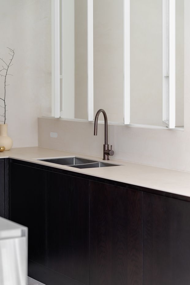 A minimalist kitchen sink area with dark timber cabinetry, a pale stone benchtop and matching splashback, a deep stainless-steel undermount sink, and a dark metal gooseneck tap. Tall white-framed windows provide soft natural light, while a simple neutral-toned ceramic vase adds understated styling.