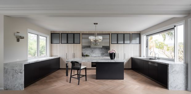 A bright, spacious contemporary kitchen viewed straight on, with a symmetrical U-shaped layout and central island. The room features dark matte cabinetry, pale timber herringbone flooring, white and grey marble-look benchtops and splashback, extensive windows, and black-framed glass-front overhead cabinets. A small breakfast setting sits beside the island, while sculptural pendant lighting and layered cornice detailing add refinement.