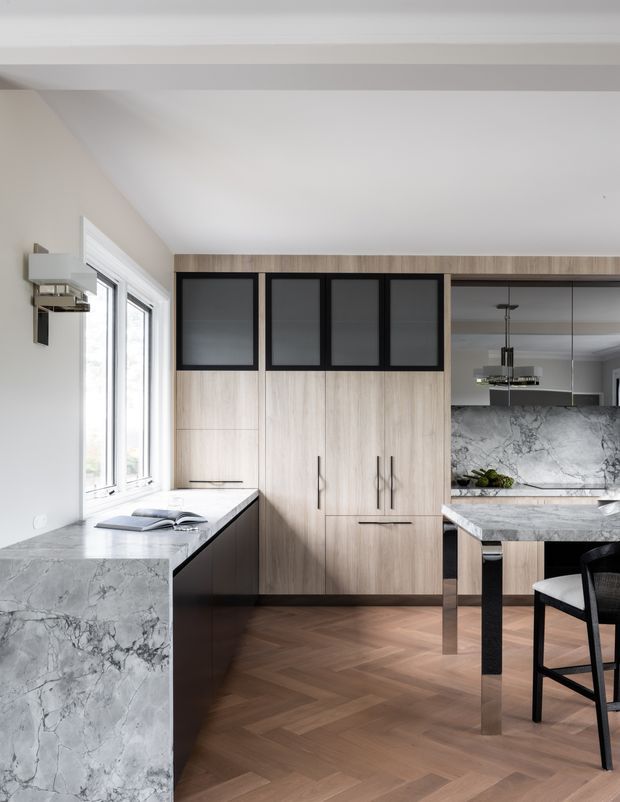 A refined contemporary kitchen with a large marble-look stone island and matching window-side benchtop. Full-height light timber cabinetry features black-framed dark glass overhead cupboards, integrated appliances, and slim black hardware. A mirrored backsplash reflects pendant lighting and additional cabinetry, while warm herringbone timber flooring and large windows create a bright, sophisticated interior.