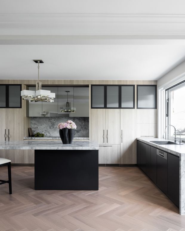 A refined contemporary kitchen with a large black island, pale timber cabinetry, dark-framed upper glass-front doors, and a dramatic grey-veined stone benchtop and splashback. The room features herringbone timber flooring, integrated appliances, pendant lighting, and bright natural light from the large window on the right.