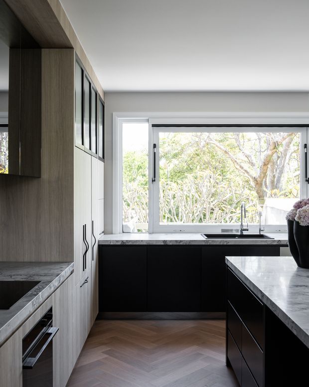 A refined contemporary kitchen with dark matte cabinetry, tall light-wood-grain storage, a large marble-look island benchtop, and a matching stone-look work surface beneath a wide garden-facing window. The floor is laid in a warm herringbone timber pattern, with black-framed glazing, integrated appliances, and a minimalist black sink and tap. Bright natural light contrasts with the dark cabinetry.