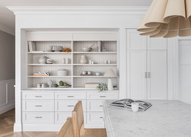 A refined white kitchen with a large marble island in the foreground, built-in cabinetry, glass-front display shelving, and paneled storage cupboards. Woven timber bar stools and a sculptural natural-fibre pendant add warmth to the otherwise light, classic interior.