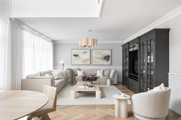 Bright, refined open-plan living and dining room with a large cream sectional sofa, boucle lounge chair, pale timber herringbone flooring, light neutral furnishings, and full-height sheer curtains. A dark charcoal built-in glazed display and media cabinet forms a strong feature wall, while framed abstract artwork, a sculptural pendant, and white wall panelling add polished contemporary detailing.