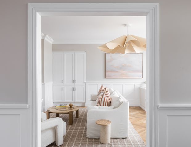 A bright, refined living room viewed through a wide, heavily moulded doorway. The room features white panelled built-in cabinetry and wall panelling, pale grey walls, herringbone timber flooring, a large textured rug, curved white seating, a round timber coffee table, a sculptural natural-toned pendant light, and soft abstract artwork. The overall scheme is light, elegant, and contemporary with traditional architectural detailing.