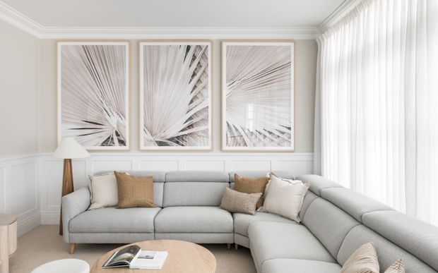Bright, refined living room featuring a large light-grey L-shaped sectional sofa, pale timber coffee table, neutral cushions, white sheer curtains, and white painted wall panelling with ornate ceiling cornices. Three framed abstract botanical artworks form a feature wall, creating a calm contemporary-classic atmosphere.