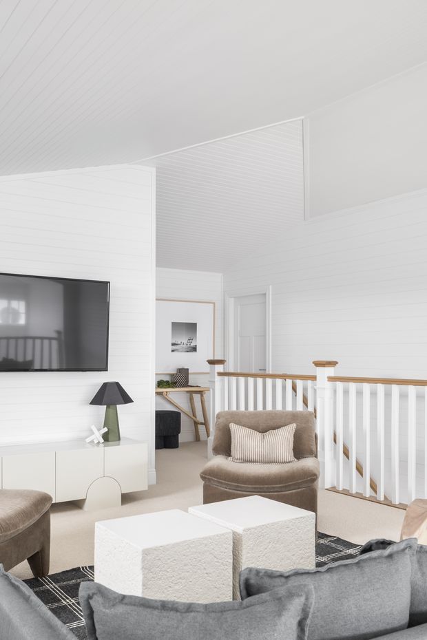 Bright, refined living room in a white-painted coastal-style interior. The room features horizontal wall panelling, a white slatted ceiling, a wall-mounted television, low white media cabinetry, neutral upholstered seating, textured white cube coffee tables, and a staircase or landing railing with white balusters and timber handrail. A small console and artwork are visible in the background.